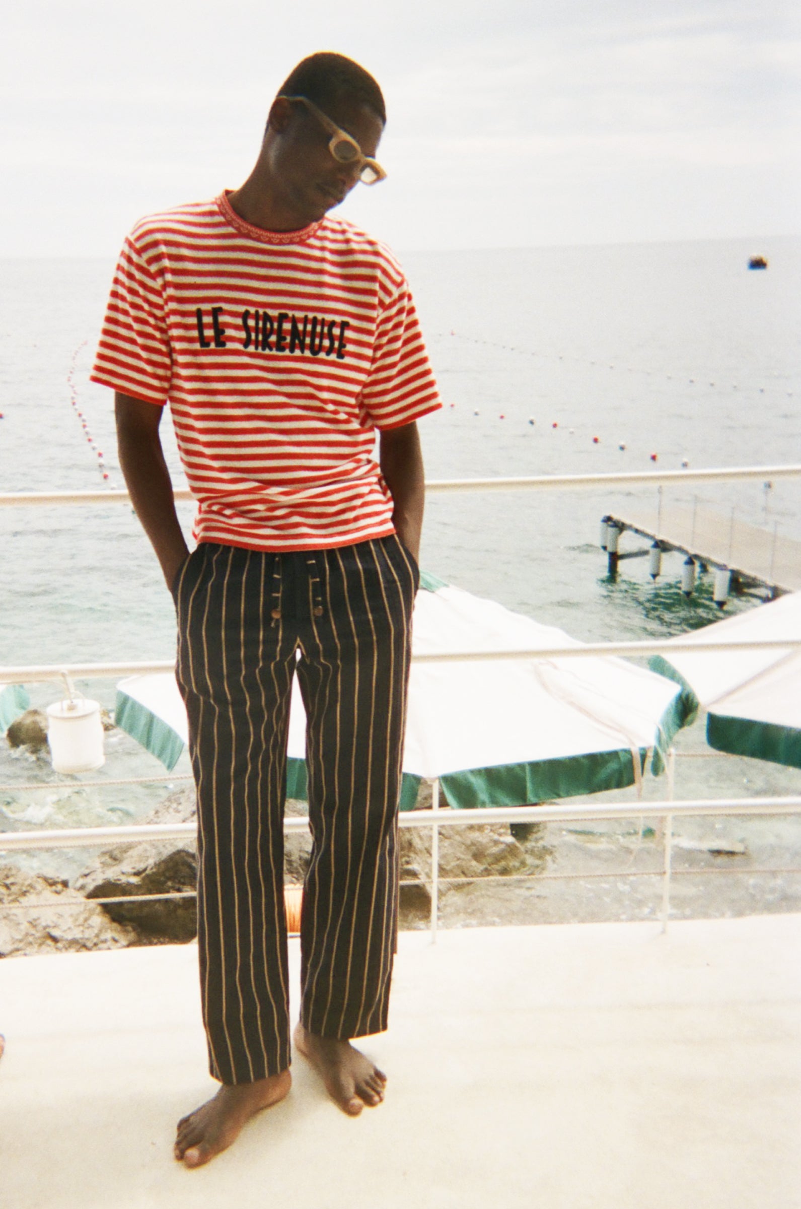 Person wearing a red and white striped shirt with 'Le Sirenuse' embroidered, and black and white striped pants standing on a deck.