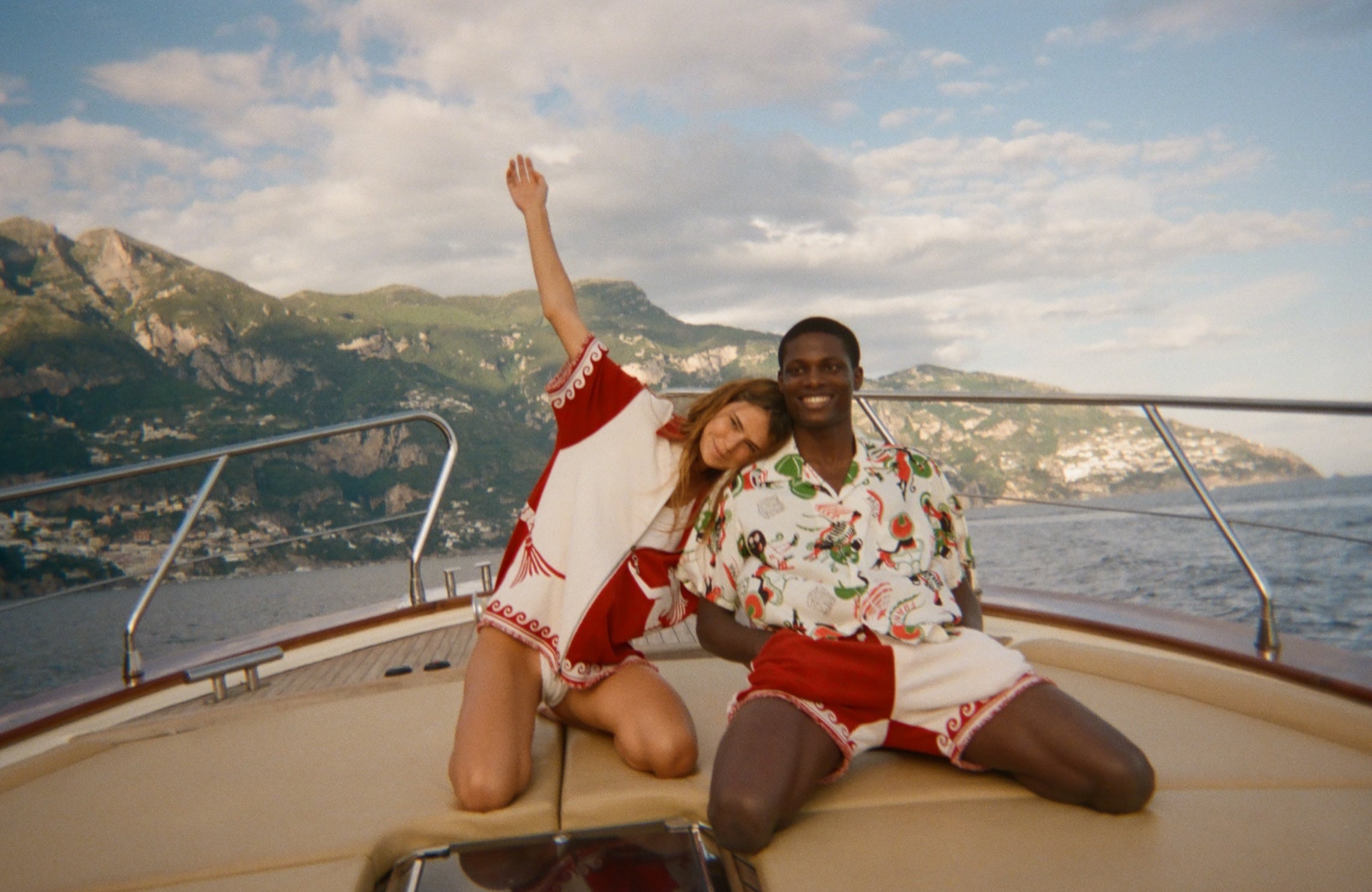 Two people sitting on a boat with scenic mountains in the background, wearing items from the Le Sirenuse x Tombolo collection.