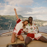 Two people sitting on a boat with scenic mountains in the background, wearing items from the Le Sirenuse x Tombolo collection.