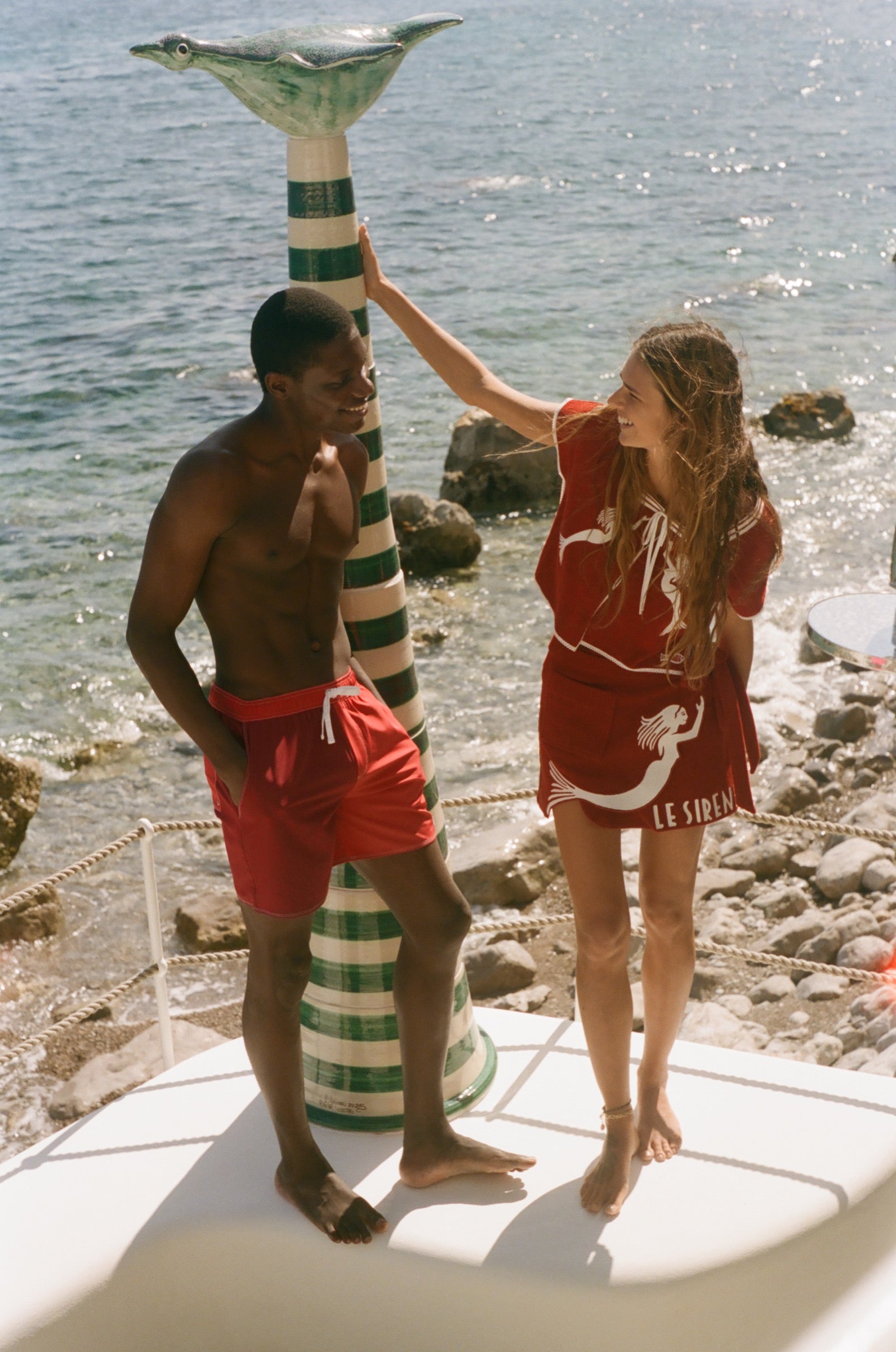 Two people in Le Sirenuse red, standing in front of a rocky beach with ocean in the background.