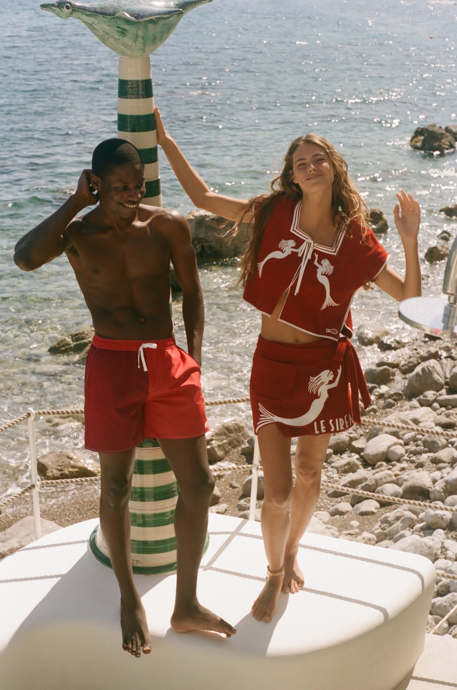 Two people in Le Sirenuse red, standing in front of a rocky beach with ocean in the background.