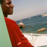 Man in a red shirt sitting by a body of water with boats and rocks.