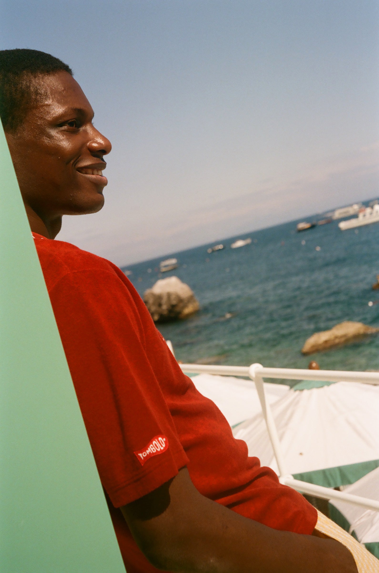 Man in a red shirt sitting by a body of water with boats and rocks.
