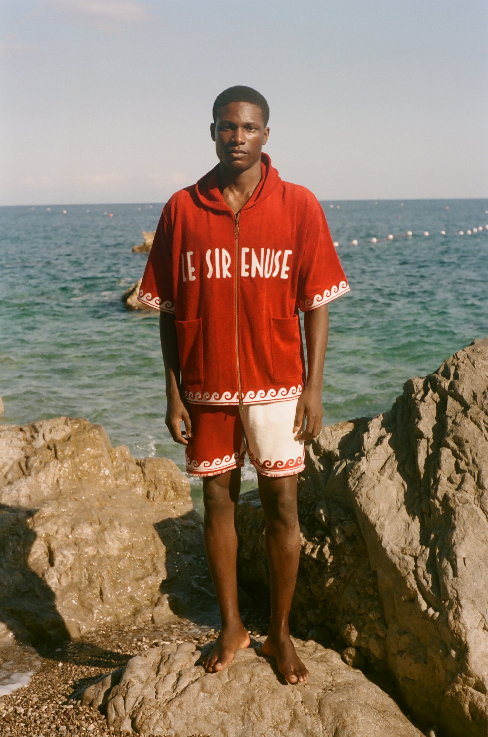Man wearing Le Sirenuse x Tombolo collection items, standing in front of the ocean. 