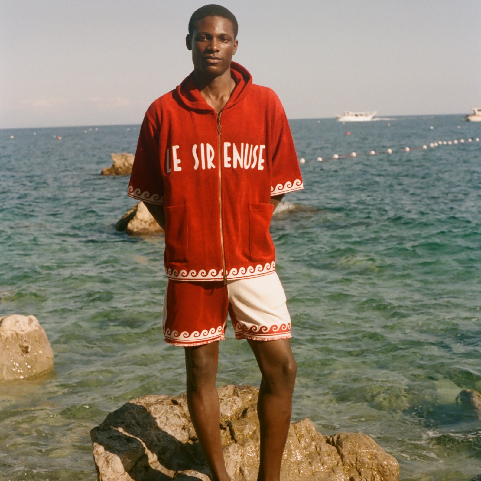 Man wearing beach towel top with matching red Le Sirenuse shorts, in front of the ocean. 