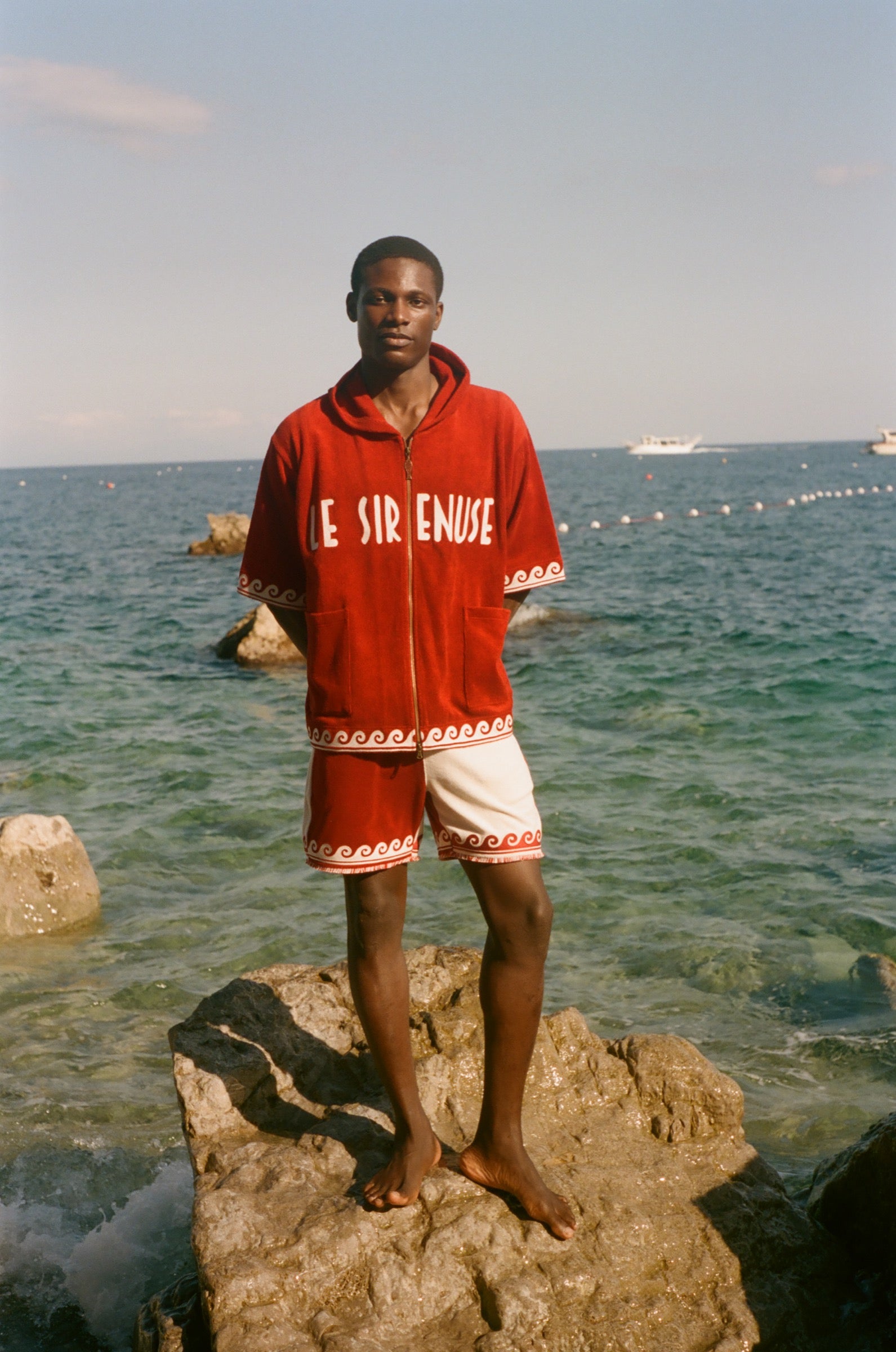 Man wearing beach towel top with matching red Le Sirenuse shorts, in front of the ocean. 