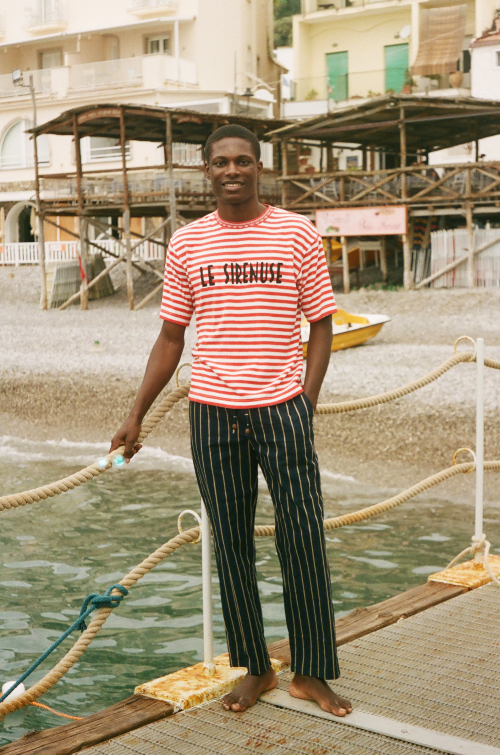 Man standing on a dock wearing a red and white Le Sirenuse striped shirt and black pants.