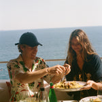 Two people enjoying a meal by the ocean with drinks and food on a table.