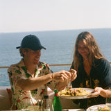 Two people enjoying a meal by the ocean with drinks and food on a table.