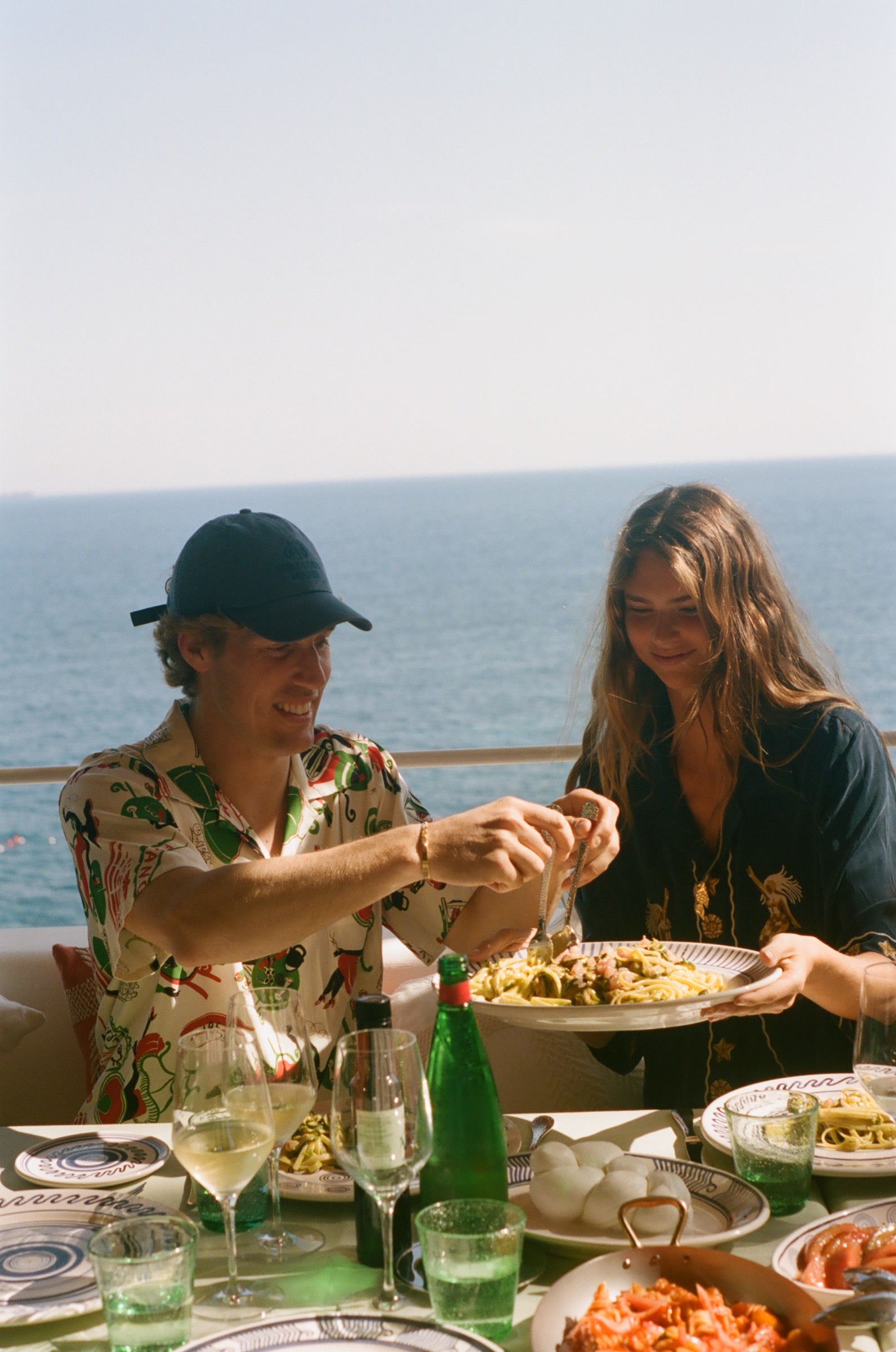 Two people enjoying a meal by the ocean with drinks and food on a table.