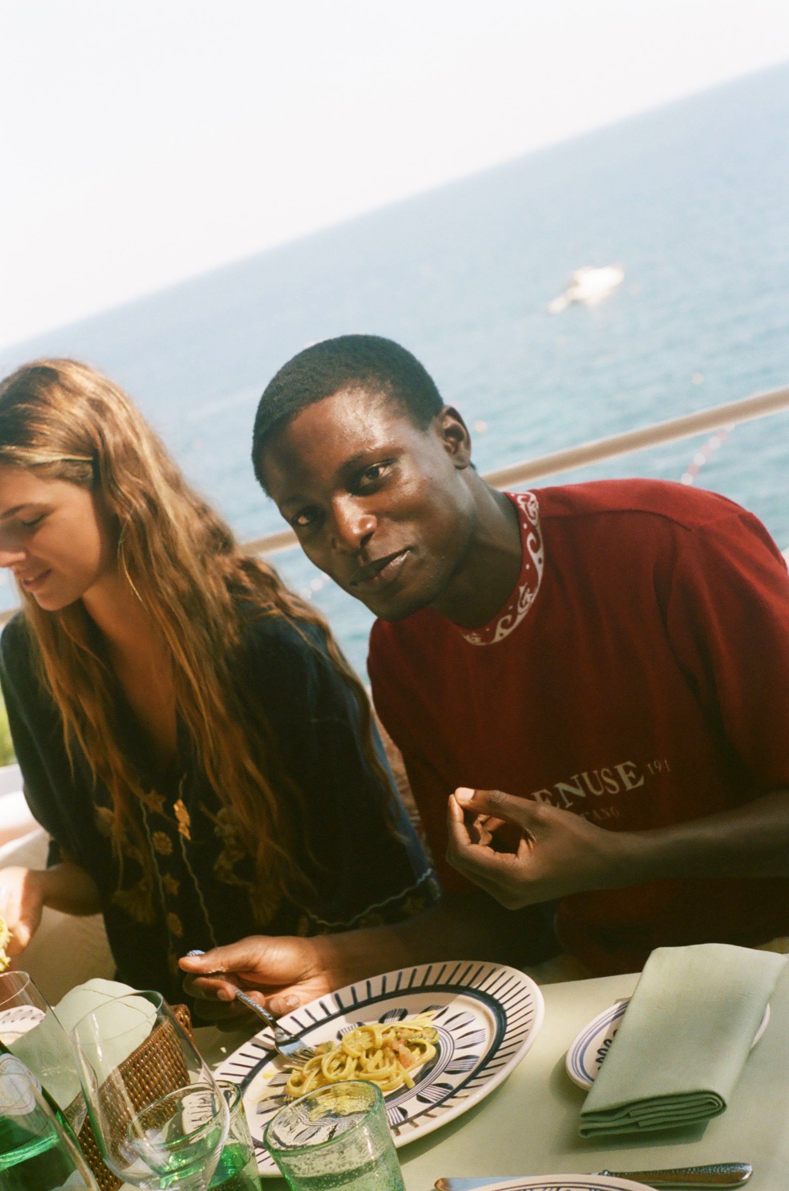 Two people sitting at a table with food and drinks outdoors, wearing the Le Sirenuse x Tombolo collaboration.