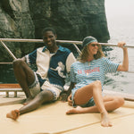 Two people sitting on a boat with a rocky cliff in the background, wearing Le Sirenuse x Tombolo merch.