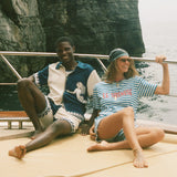 Two people sitting on a boat with a rocky cliff in the background, wearing Le Sirenuse x Tombolo merch.