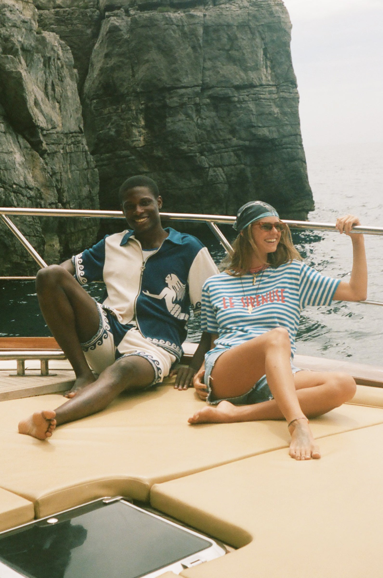 Two people sitting on a boat with a rocky cliff in the background, wearing Le Sirenuse x Tombolo merch.