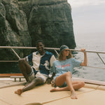 Two people sitting on a boat with cliffs and ocean in the background, wearing Le Sirenuse x Tombolo collection items.