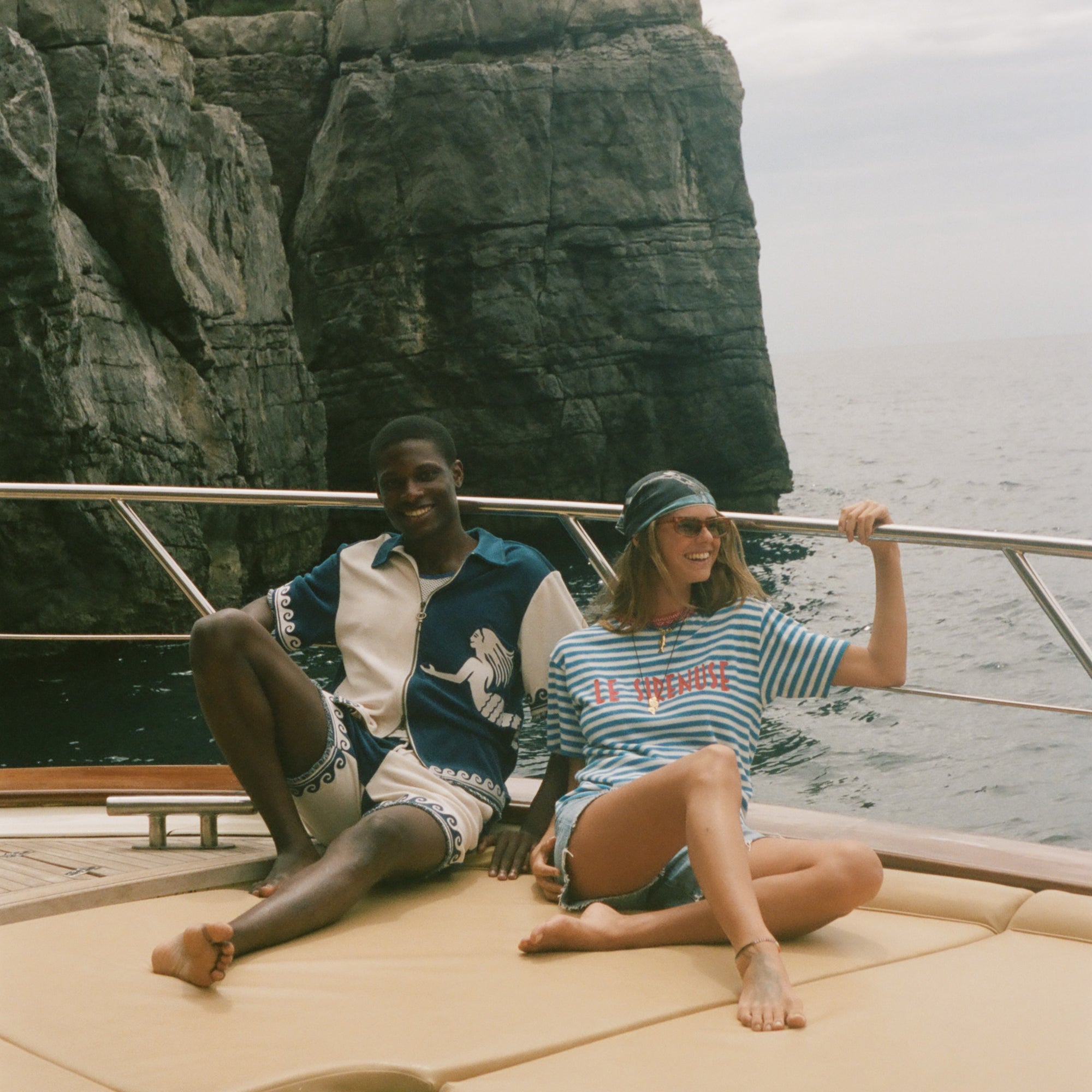 Two people sitting on a boat with cliffs and ocean in the background, wearing Le Sirenuse x Tombolo collection items.
