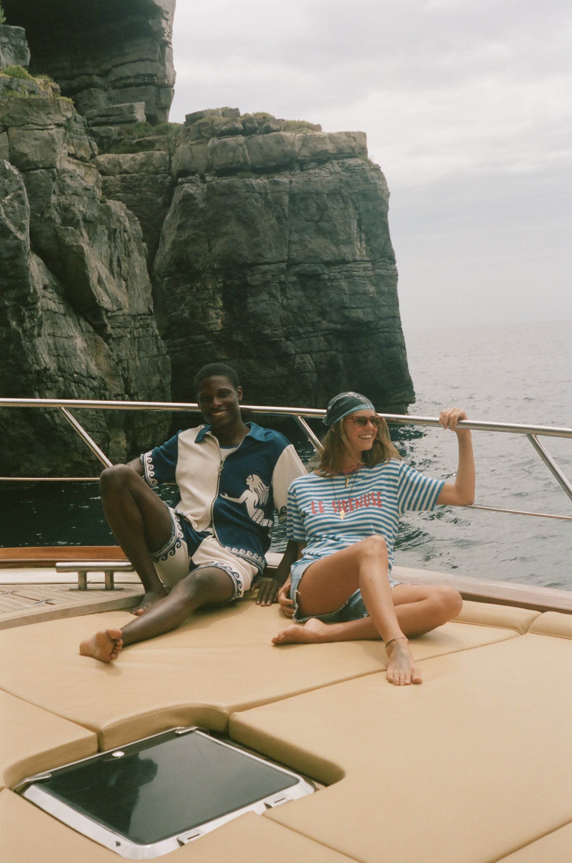 Two people sitting on a boat with cliffs and ocean in the background, wearing Le Sirenuse x Tombolo collection items.