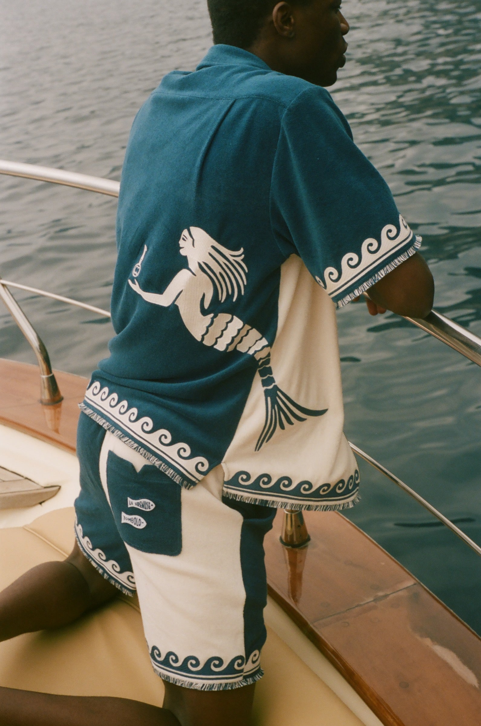 Man wearing Blue Late checkout set, sitting on the edge of a boat. 