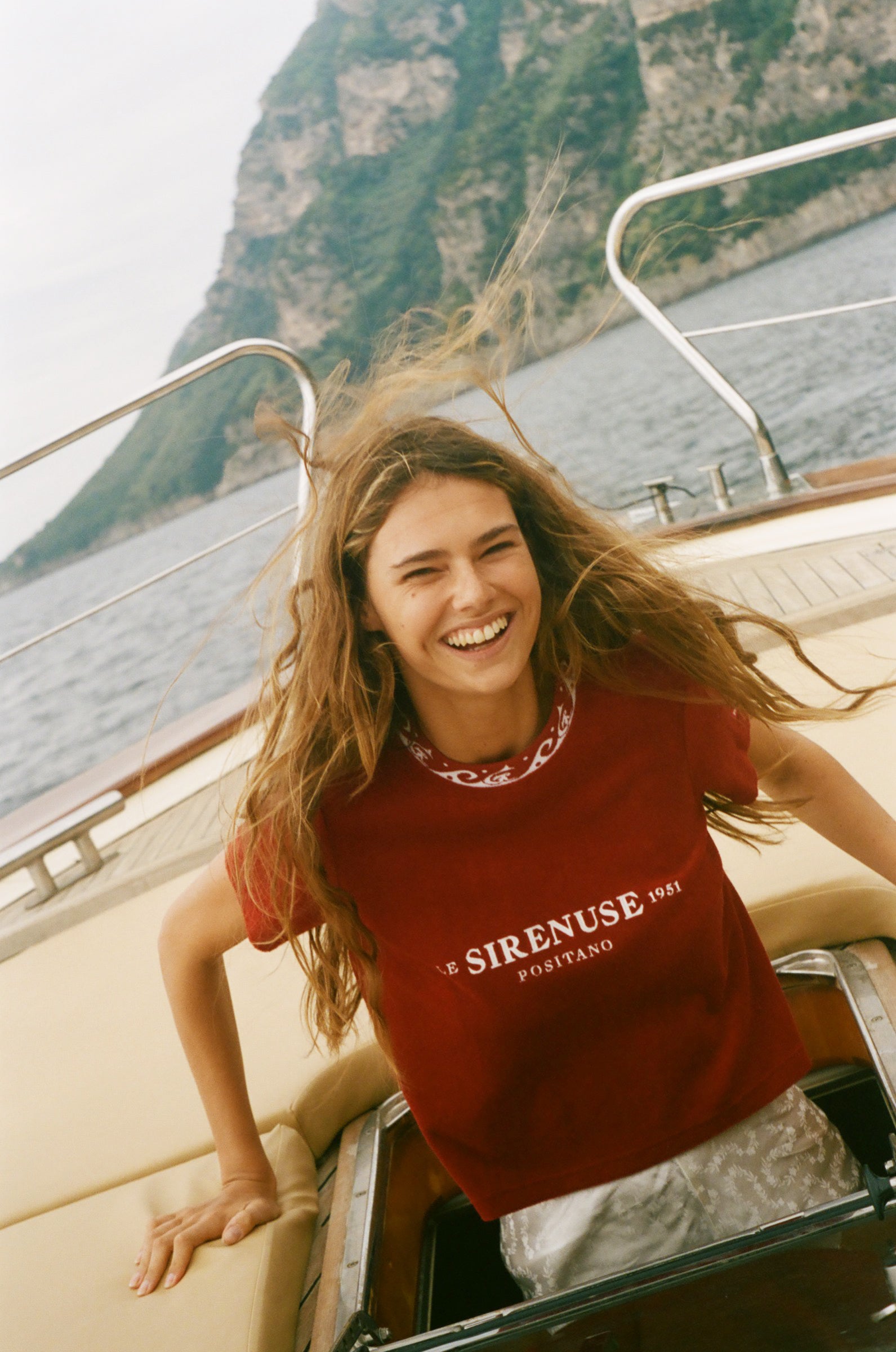 Woman in a red 'Sirenuse' t-shirt on a boat with a mountainous background