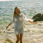 Image of woman laughing as she wades out of ankle deep ocean, wearing a light blue sand dollar bikini shirt and matching skirt. 