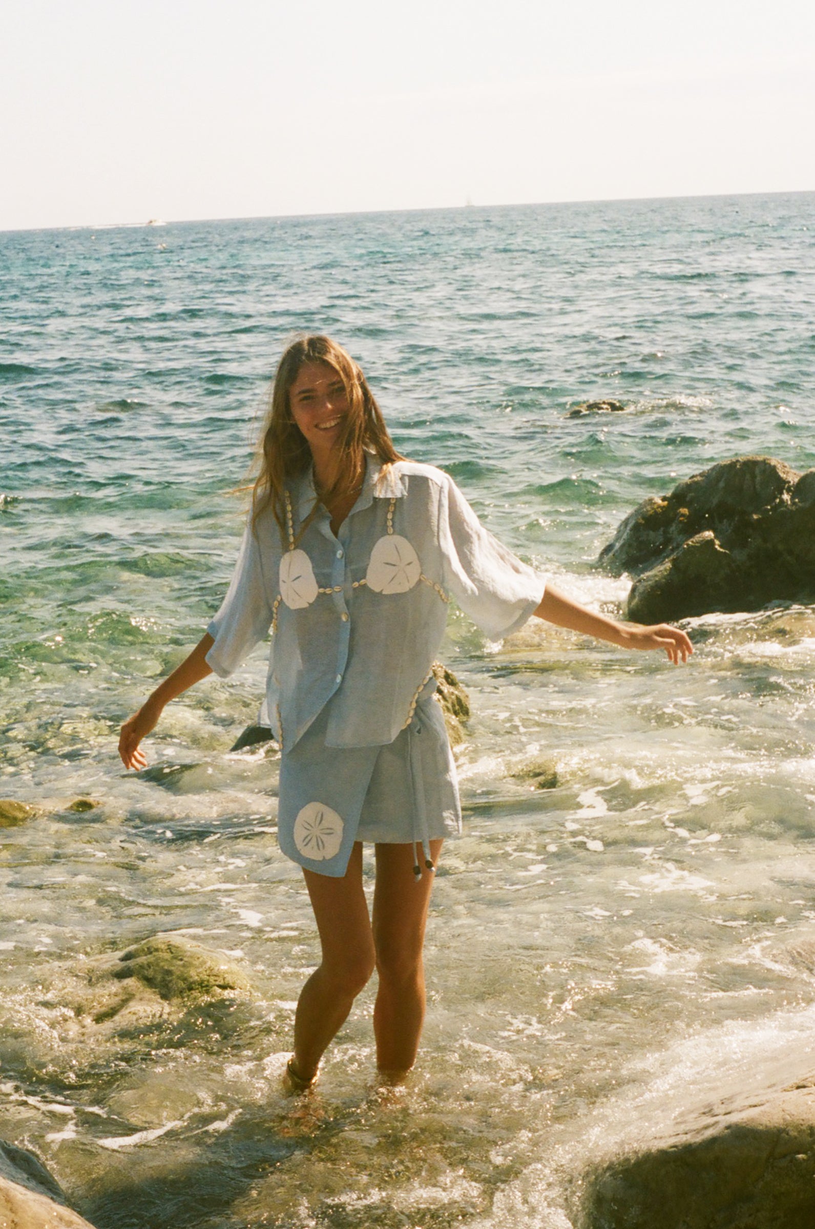 Image of woman laughing as she wades out of ankle deep ocean, wearing a light blue sand dollar bikini shirt and matching skirt. 