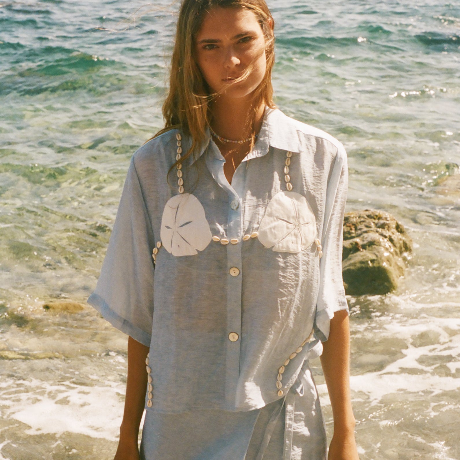 Image of woman in light blue sand dollar bikini shirt, standing ankle deep in the ocean, with a matching skirt underneath.