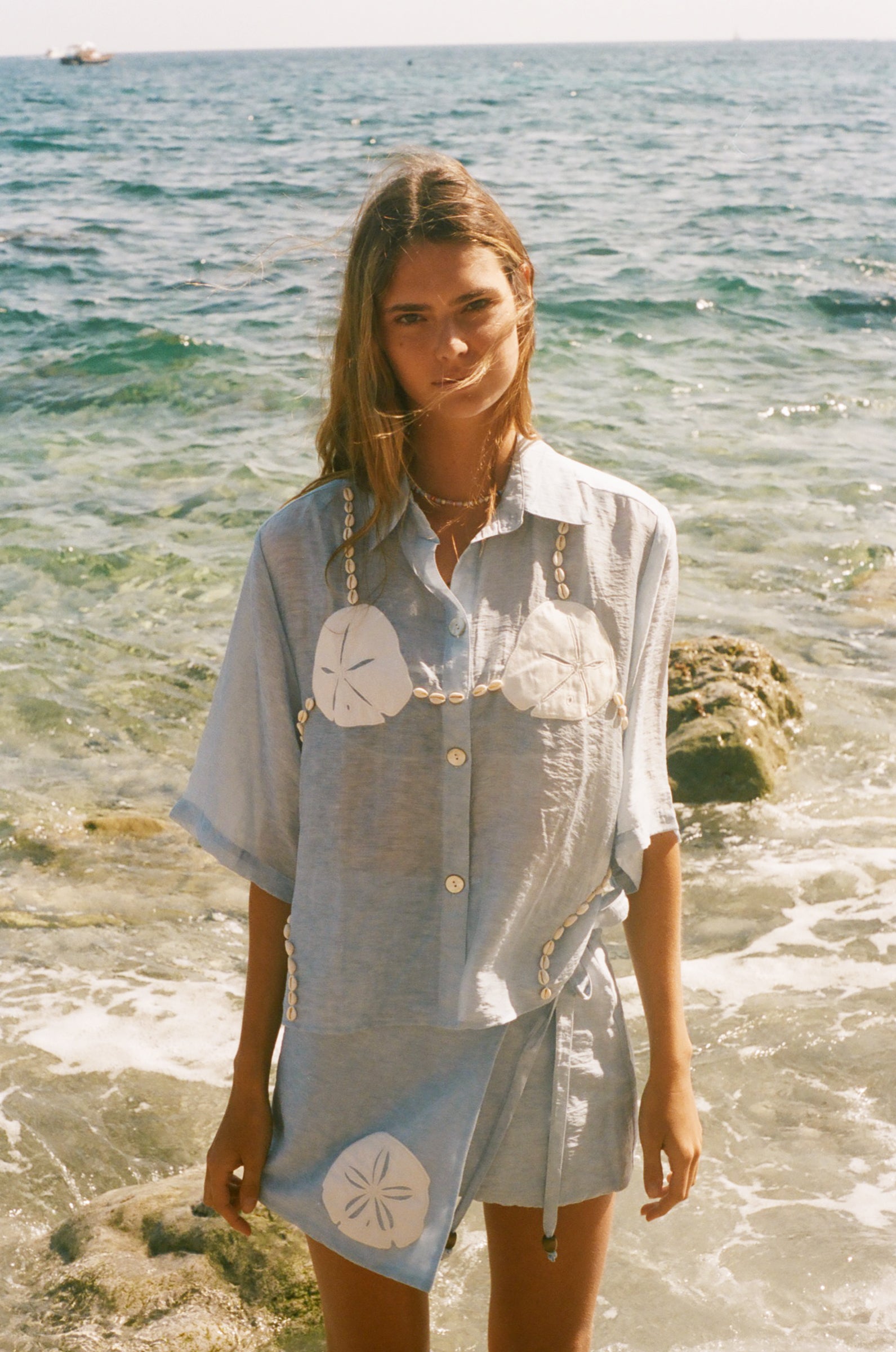 Image of woman in light blue sand dollar bikini shirt, standing ankle deep in the ocean, with a matching skirt underneath.