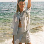 Image of woman holding her hair wearing a light blue sand dollar bikini shirt, standing at the edge of the ocean, with a matching skirt underneath.