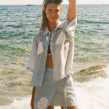 Image of woman holding her hair wearing a light blue sand dollar bikini shirt, standing at the edge of the ocean, with a matching skirt underneath.