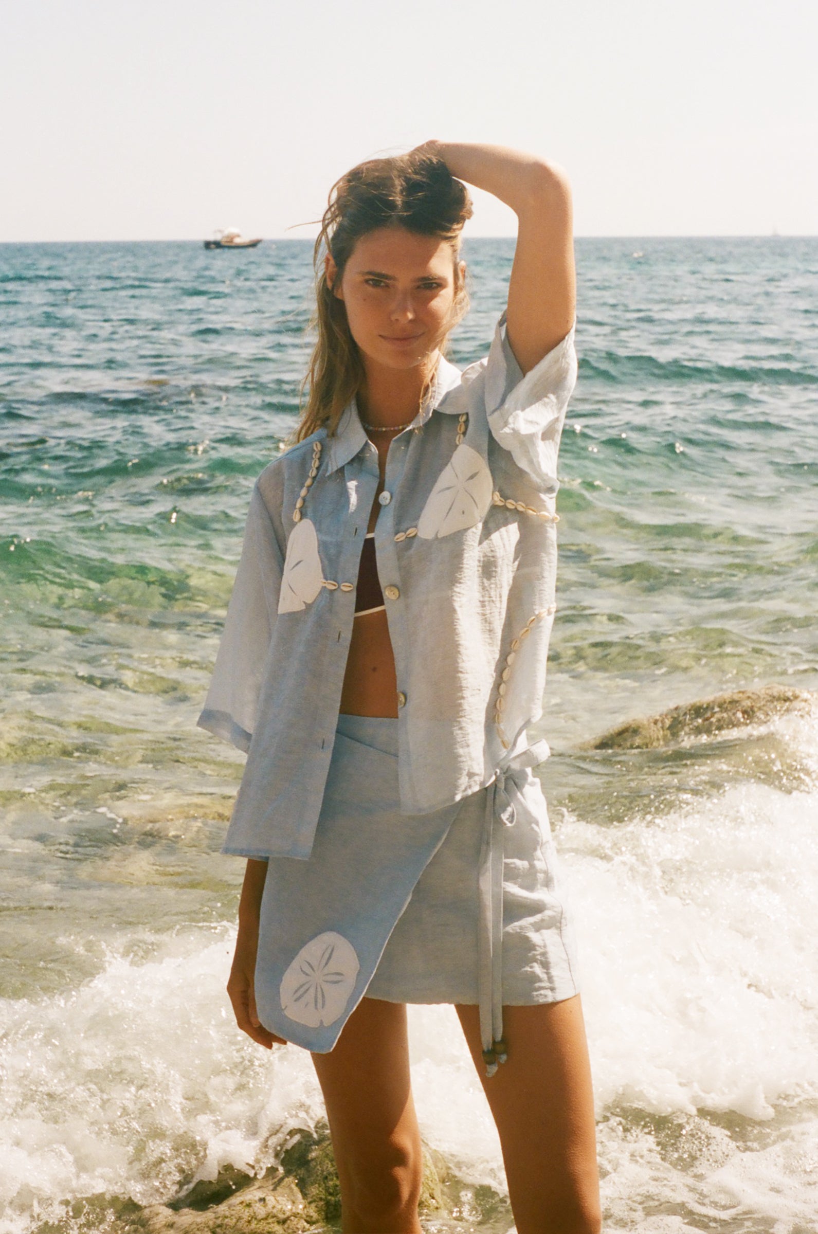 Image of woman holding her hair wearing a light blue sand dollar bikini shirt, standing at the edge of the ocean, with a matching skirt underneath.