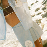 Close cropped image of woman dancing by the ocean. She is wearing a light blue sand dollar bikini shirt and matching skirt, standing by the ocean, with a maroon bikini underneath.