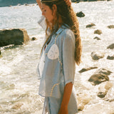 Image of woman holding her hair up, wearing a light blue sand dollar bikini shirt and matching skirt, standing by the ocean.