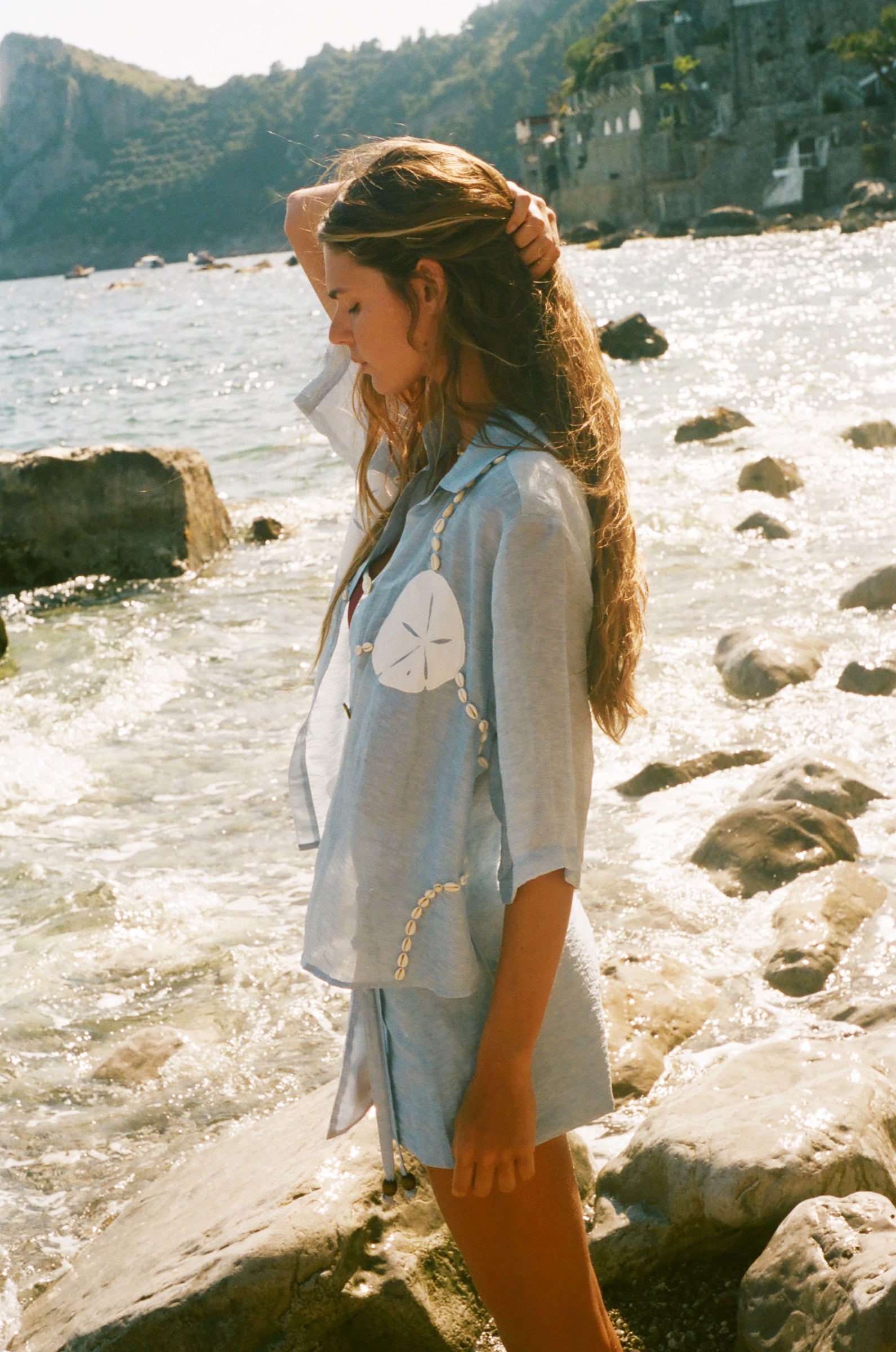 Image of woman holding her hair up, wearing a light blue sand dollar bikini shirt and matching skirt, standing by the ocean.
