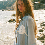 Image of woman in light blue sand dollar bikini shirt, standing at the beach shore.