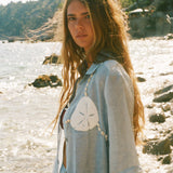Image of woman in light blue sand dollar bikini shirt, standing at the beach shore.