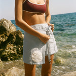Image of woman tying the knot on a light blue skirt featuring an appliquéd sand dollar, standing by the ocean, wearing a maroon bikini.