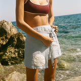 Image of woman tying the knot on a light blue skirt featuring an appliquéd sand dollar, standing by the ocean, wearing a maroon bikini.