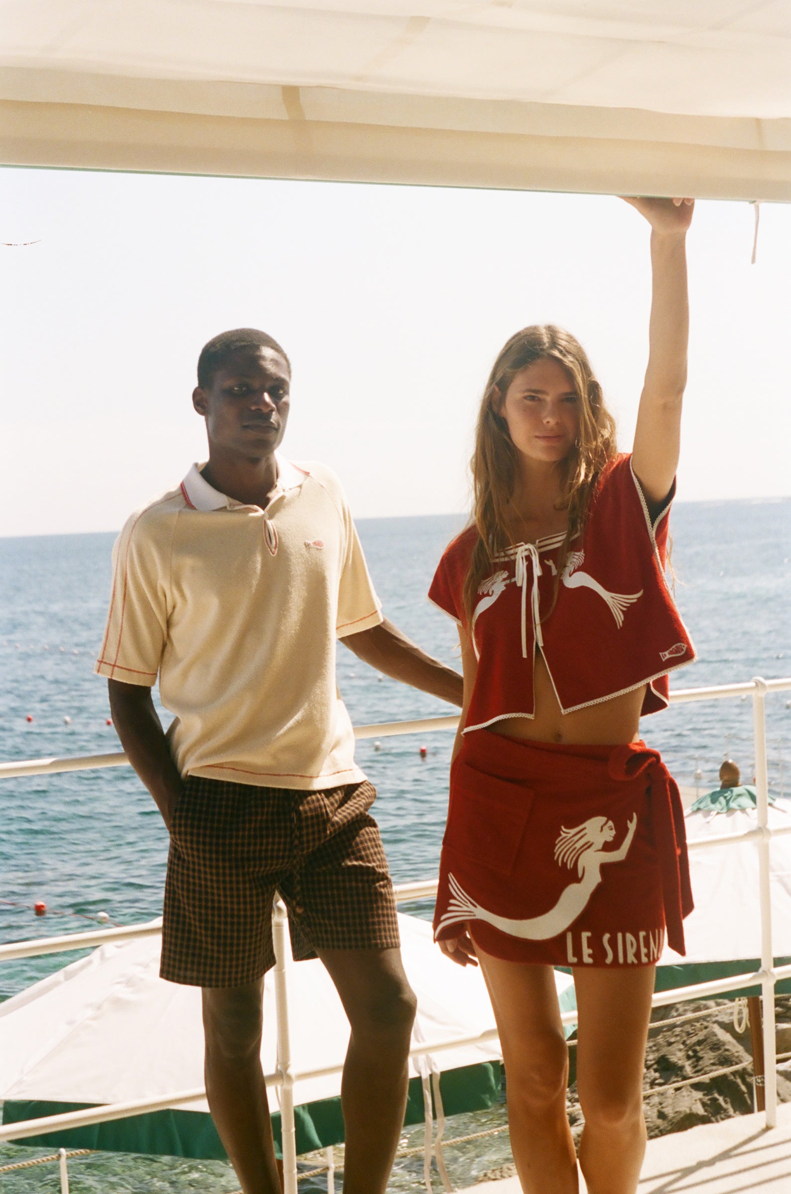 Two people wearing Le Sirenuse x Tombolo pieces, standing in front of the ocean. 