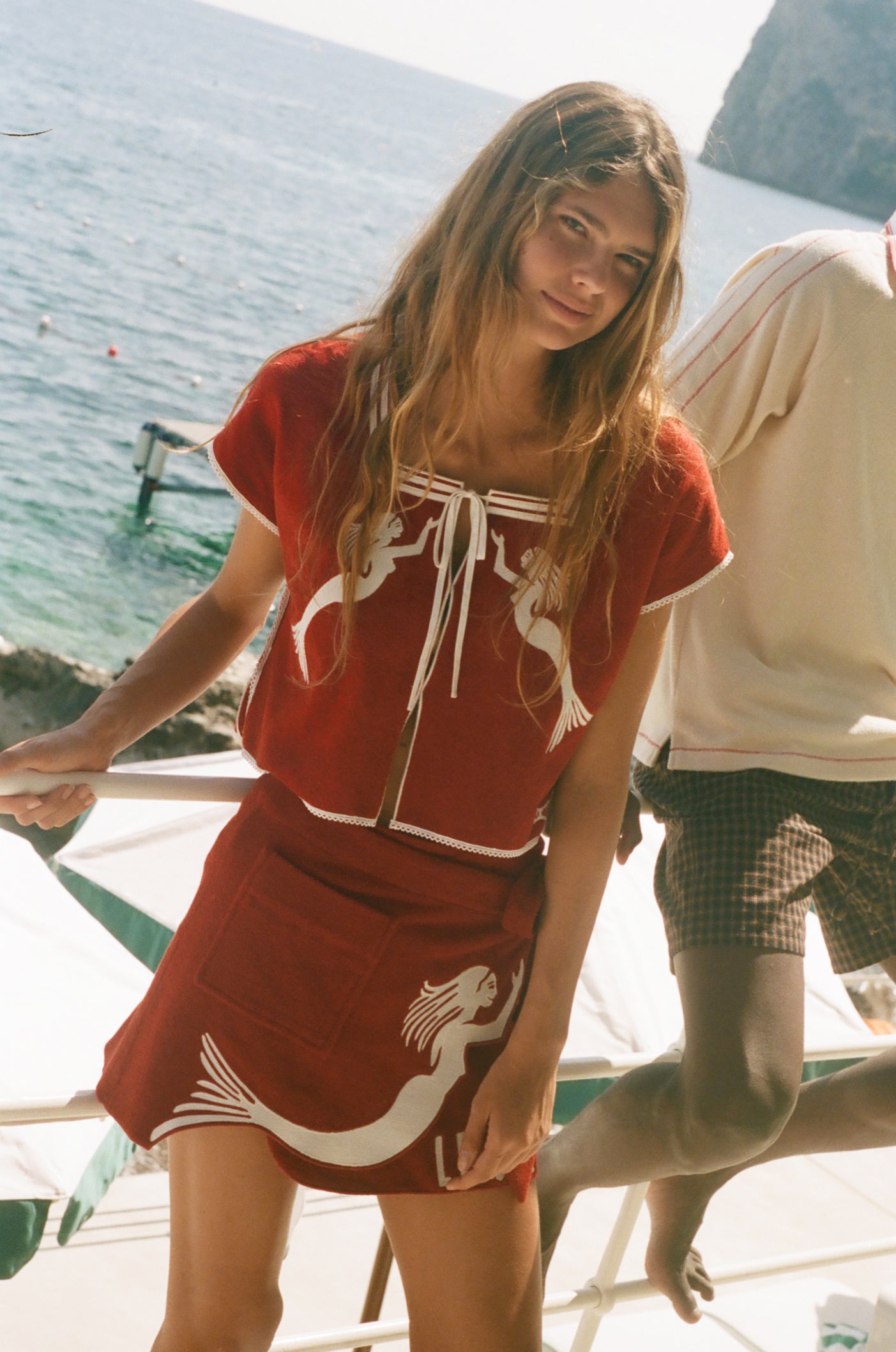 Woman wearing matching Le Sirenuse hand towel set, standing in front of the ocean. 