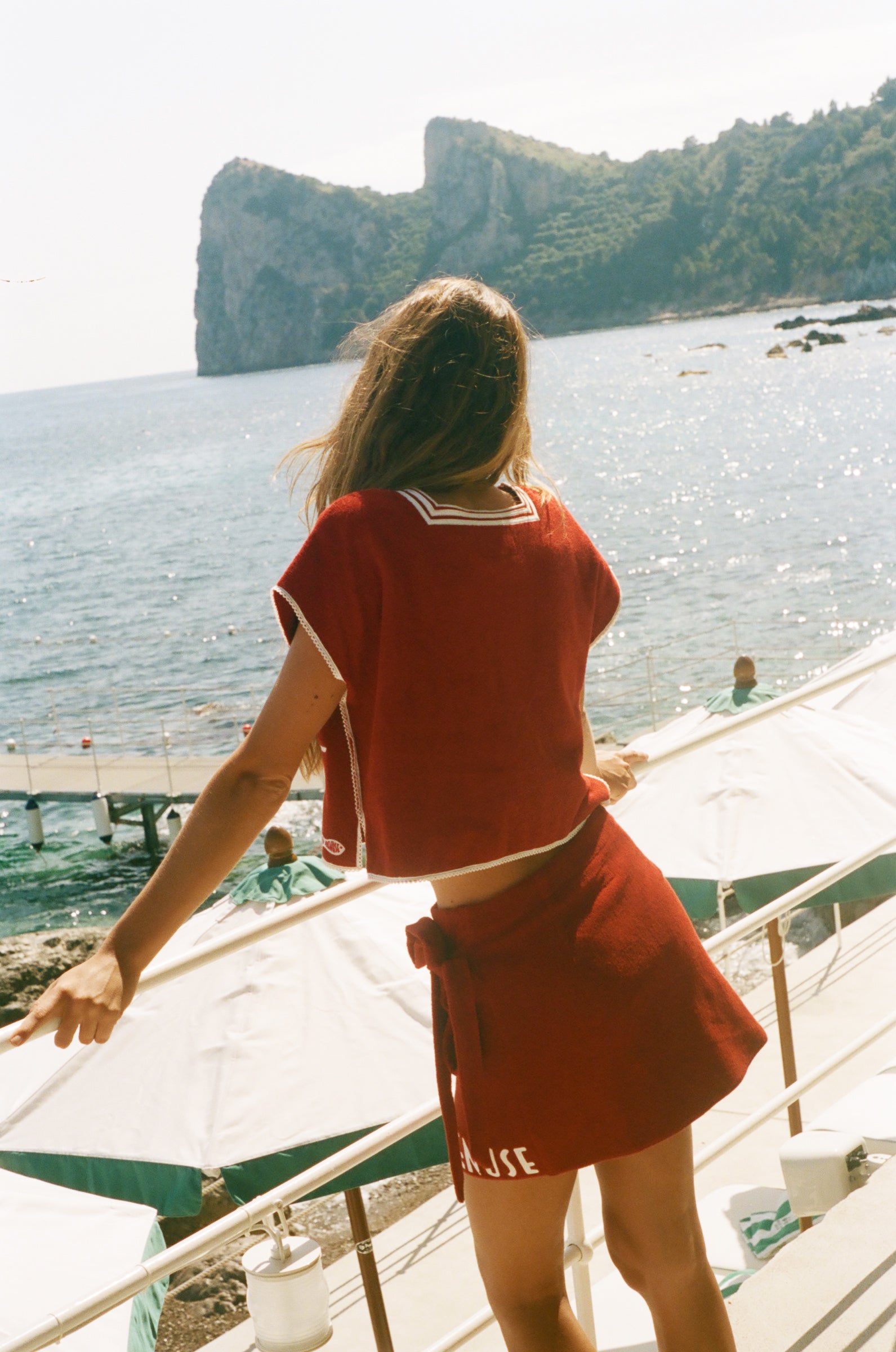 Person in a red Le Sirenuse outfit standing on a boat looking at a scenic view of water and cliffs.