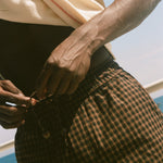 A close-up lifestyle shot of a person outdoors by the water, wearing brown and black checkered plaid shorts and a cream-colored t-shirt.