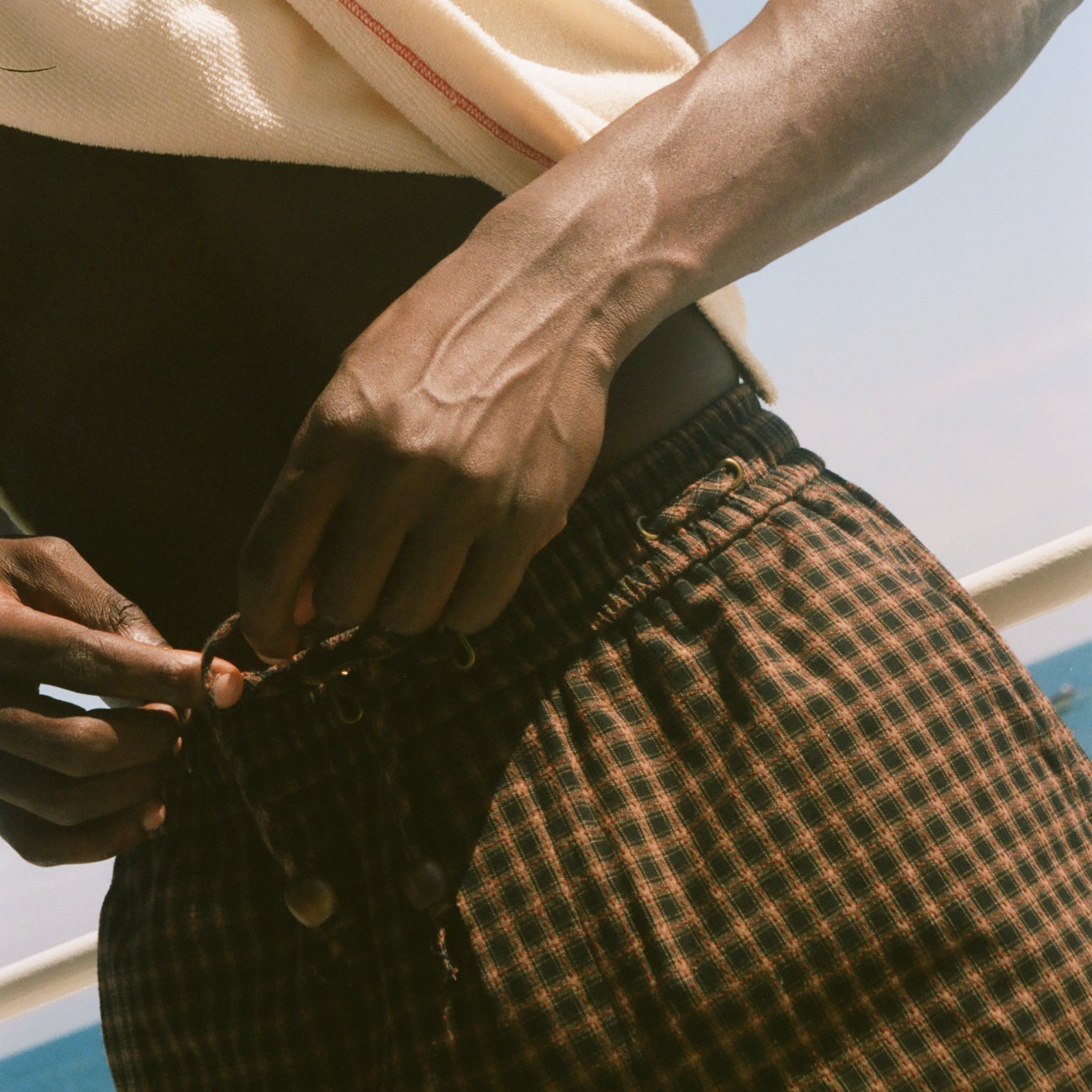 A close-up lifestyle shot of a person outdoors by the water, wearing brown and black checkered plaid shorts and a cream-colored t-shirt.