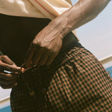 A close-up lifestyle shot of a person outdoors by the water, wearing brown and black checkered plaid shorts and a cream-colored t-shirt.