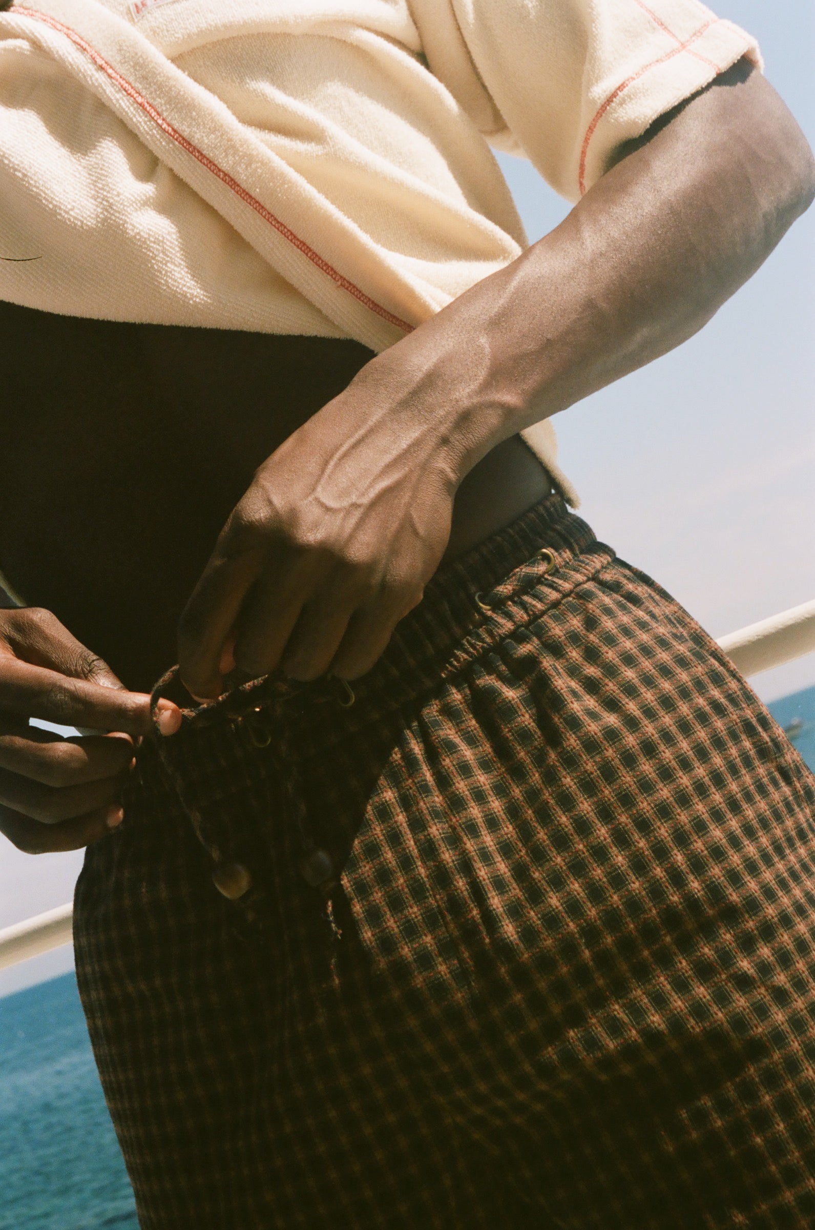 A close-up lifestyle shot of a person outdoors by the water, wearing brown and black checkered plaid shorts and a cream-colored t-shirt.