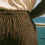 Close-up of a person wearing brown and black plaid shorts with an elastic waistband, paired with a cream knit top, with a blue ocean background visible.