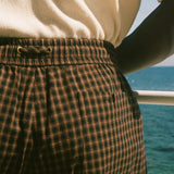 Close-up of a person wearing brown and black plaid shorts with an elastic waistband, paired with a cream knit top, with a blue ocean background visible.