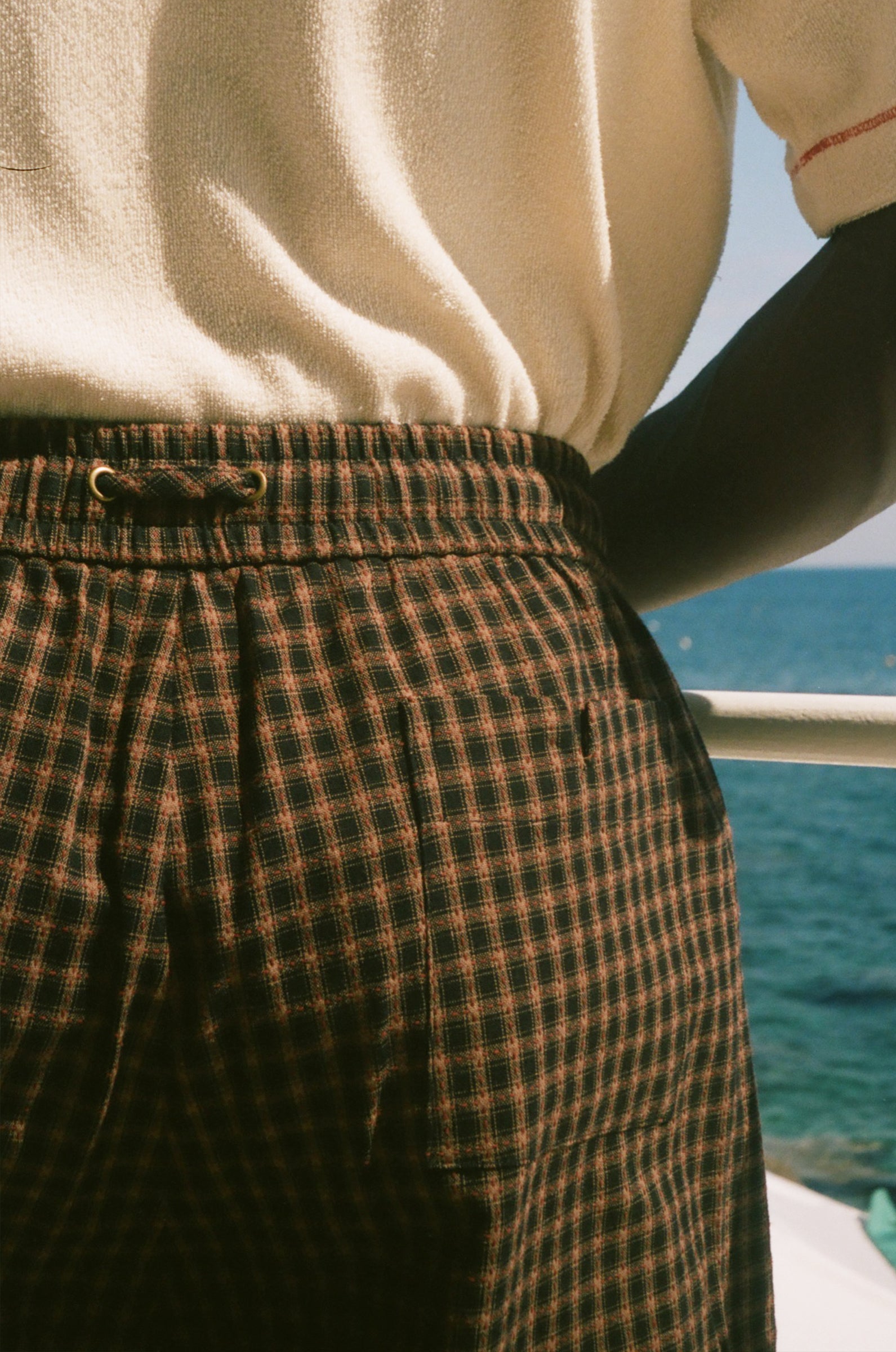 Close-up of a person wearing brown and black plaid shorts with an elastic waistband, paired with a cream knit top, with a blue ocean background visible.