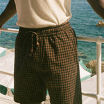 A person wearing dark brown and black checkered plaid shorts with a drawstring waist, paired with a cream-colored t-shirt, standing on a deck with the ocean in the background.