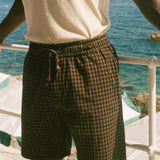 A person wearing dark brown and black checkered plaid shorts with a drawstring waist, paired with a cream-colored t-shirt, standing on a deck with the ocean in the background.