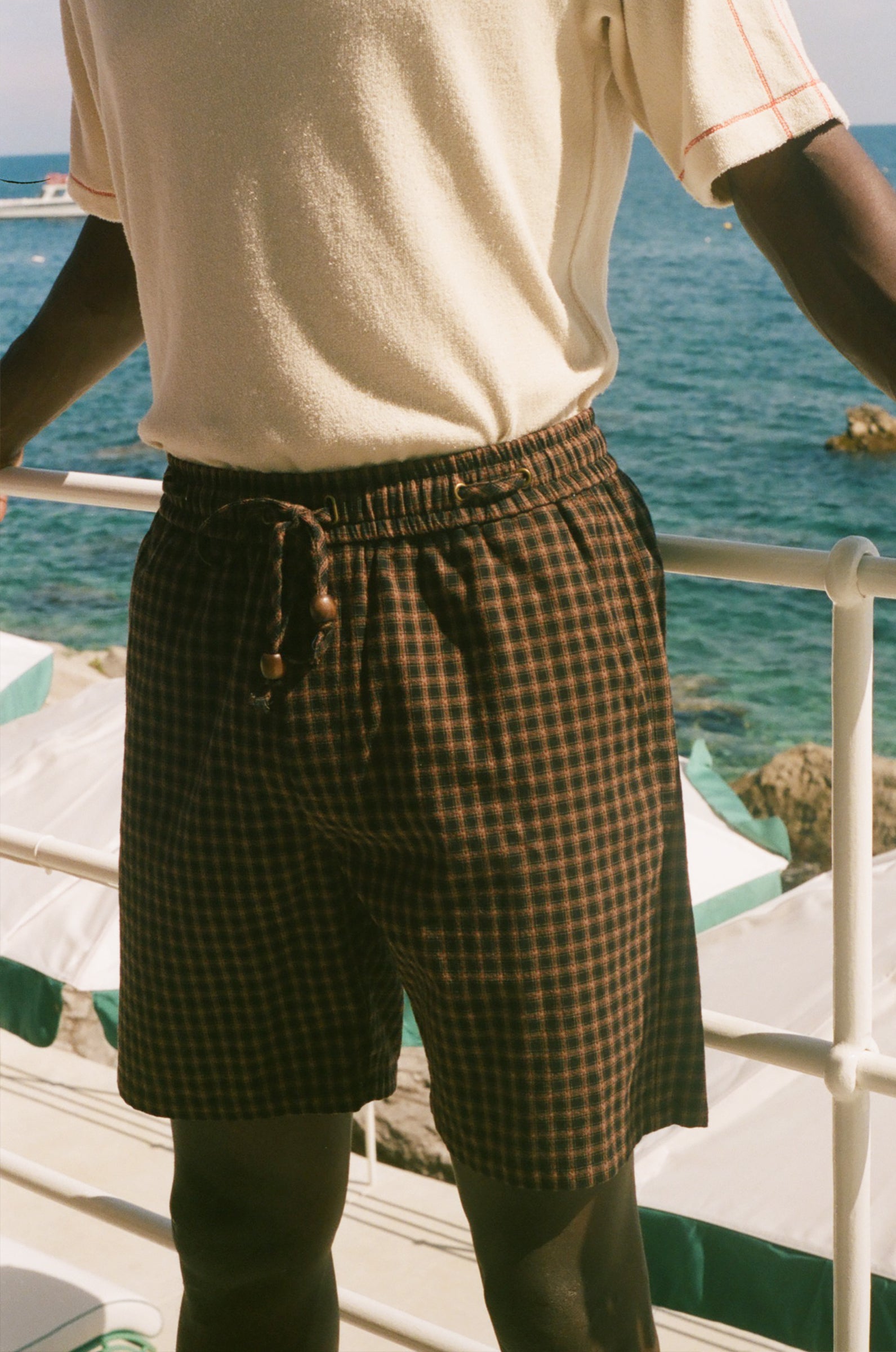 A person wearing dark brown and black checkered plaid shorts with a drawstring waist, paired with a cream-colored t-shirt, standing on a deck with the ocean in the background.